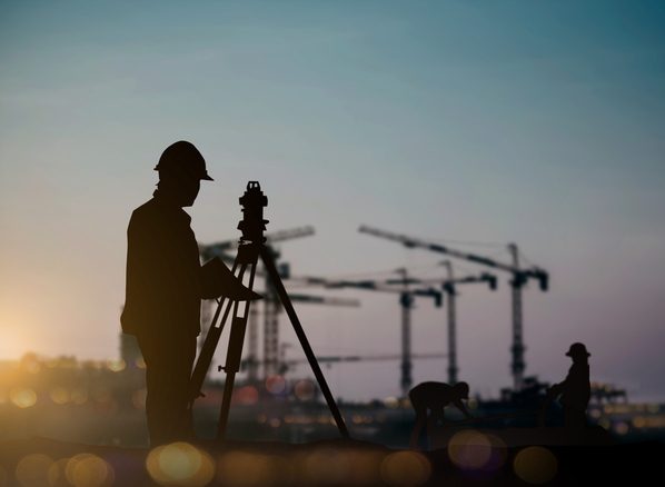 A surveyor setting up a level on a construction site at dusk, tower cranes in the background